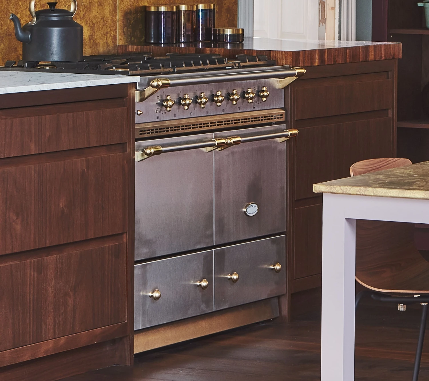 A modern kitchen featuring a stainless steel oven with brass accents, set between dark wood cabinets. A black teapot is on the marble countertop above the oven. Various jars are on the counter in the background.