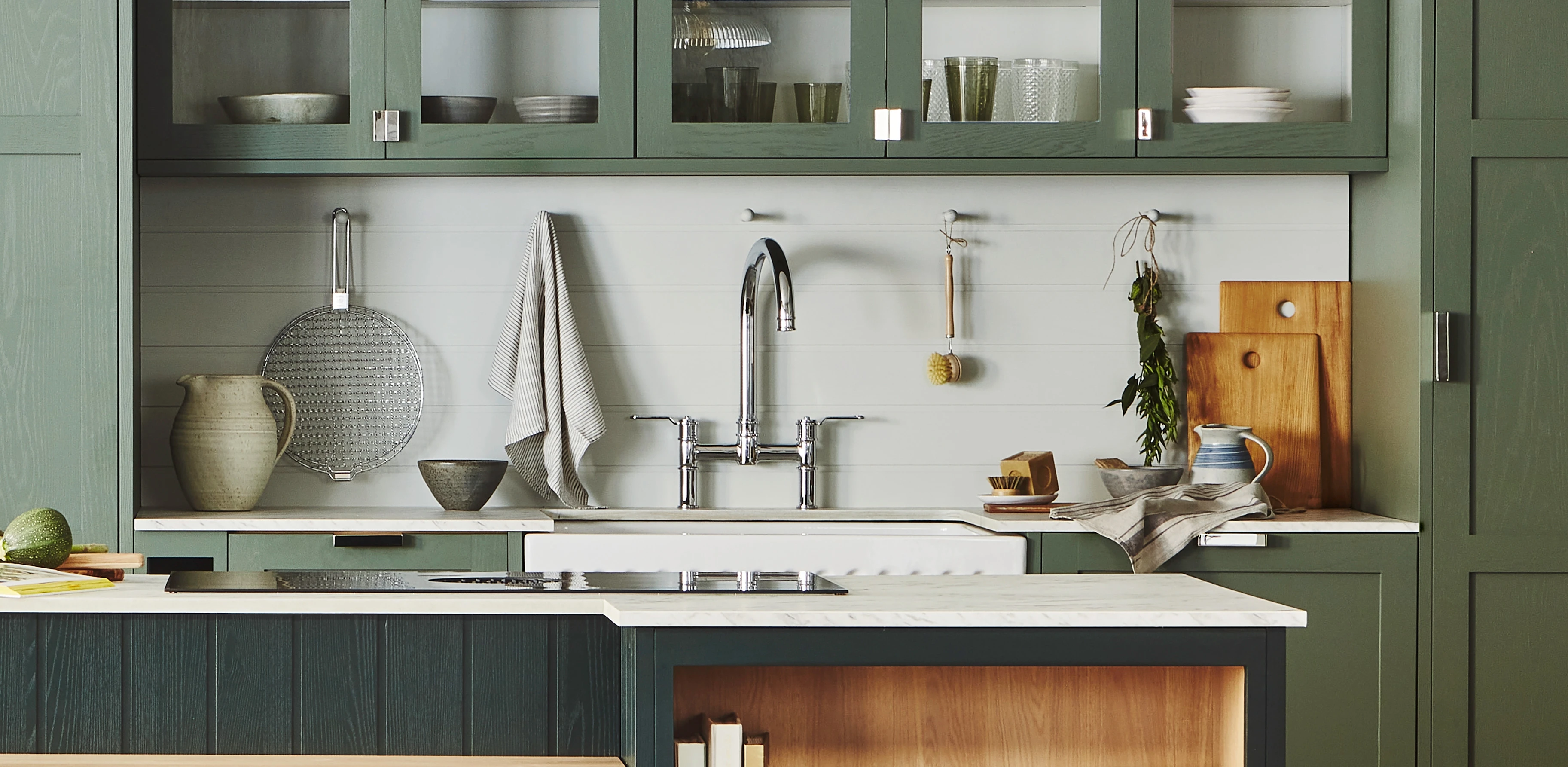 A modern kitchen with green cabinetry, white walls, and a farmhouse sink. The countertop has a cutting board, pitcher, and two bowls. Shelves display glassware and dishes. A towel hangs by the sink, and a plant adds a touch of nature.