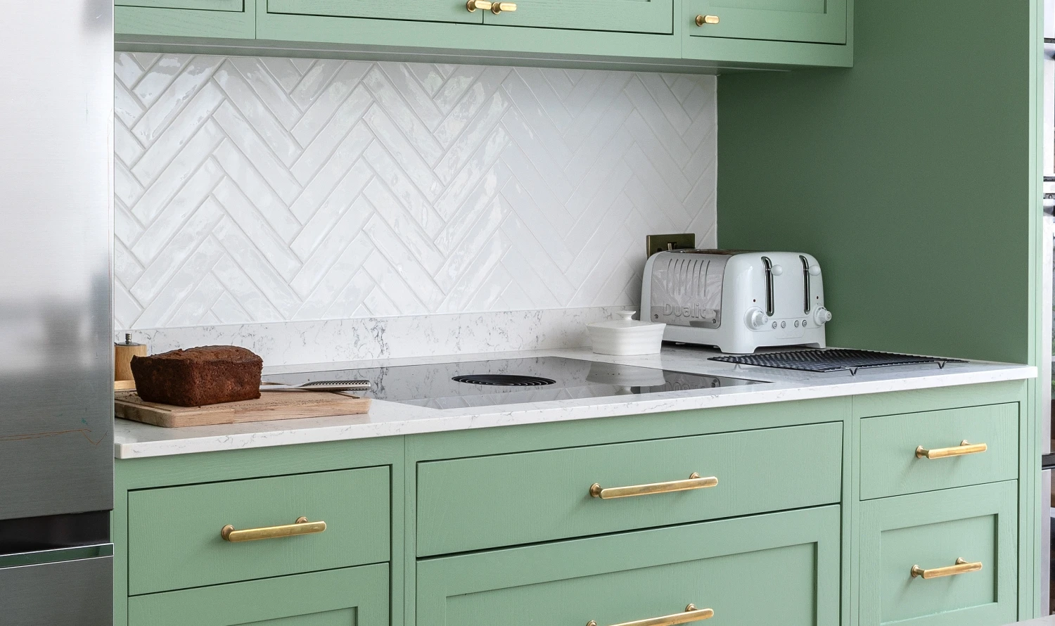 A modern kitchen with green cabinets, brass handles, and a white herringbone tile backsplash. A loaf of bread sits on a wooden cutting board on the counter next to a toaster and a stovetop.