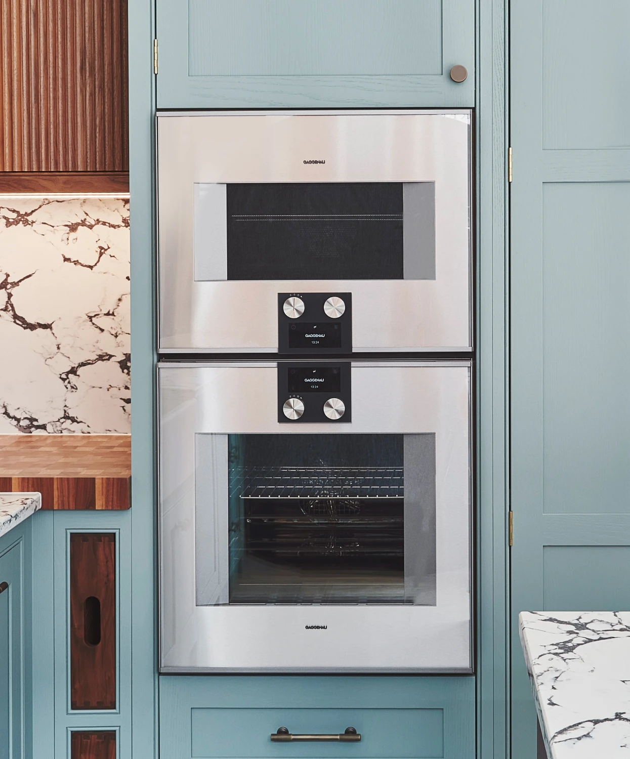 A modern kitchen with a built-in double oven set into light blue cabinetry. The oven has a stainless steel finish and digital controls. The countertop features a white and brown marble design, and there is wood paneling on the adjacent wall.