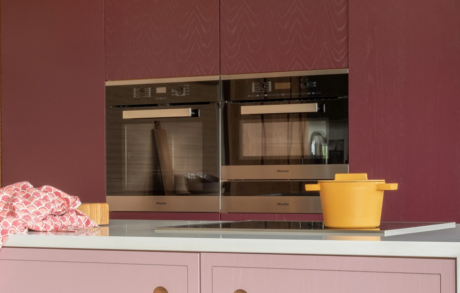 A modern kitchen with maroon cabinets, featuring two built-in ovens. A yellow pot sits on the countertop, next to a folded pink patterned cloth. The overall design is sleek and contemporary.