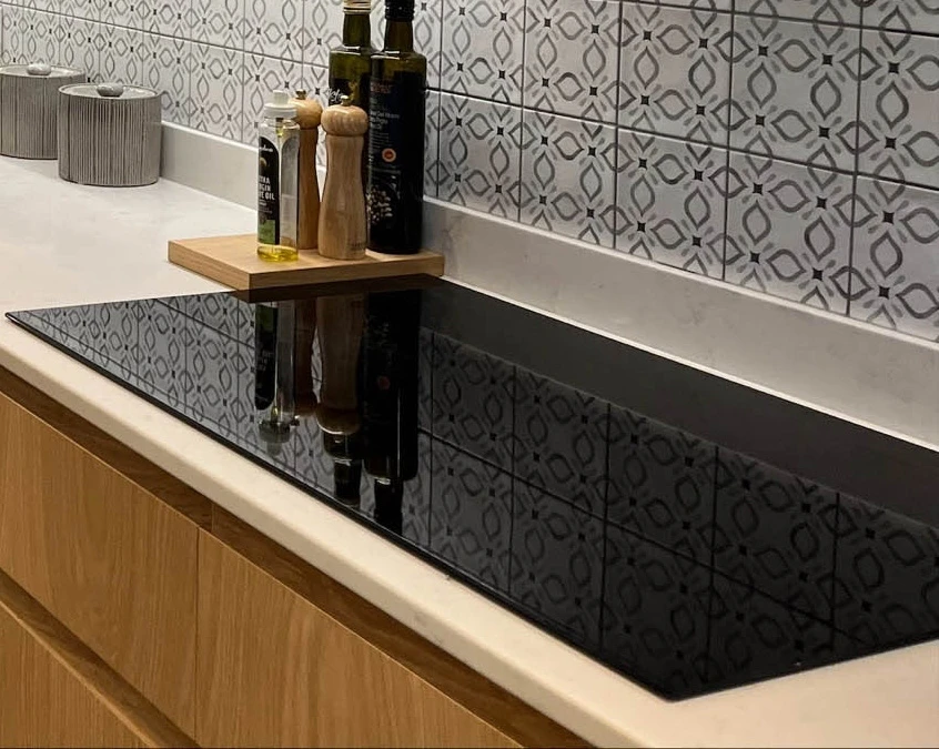 Modern kitchen counter featuring a black induction cooktop. Behind are bottles of olive oil and a wooden tray with salt and pepper shakers. The backsplash has a geometric tile pattern, and the cabinetry is light wood.