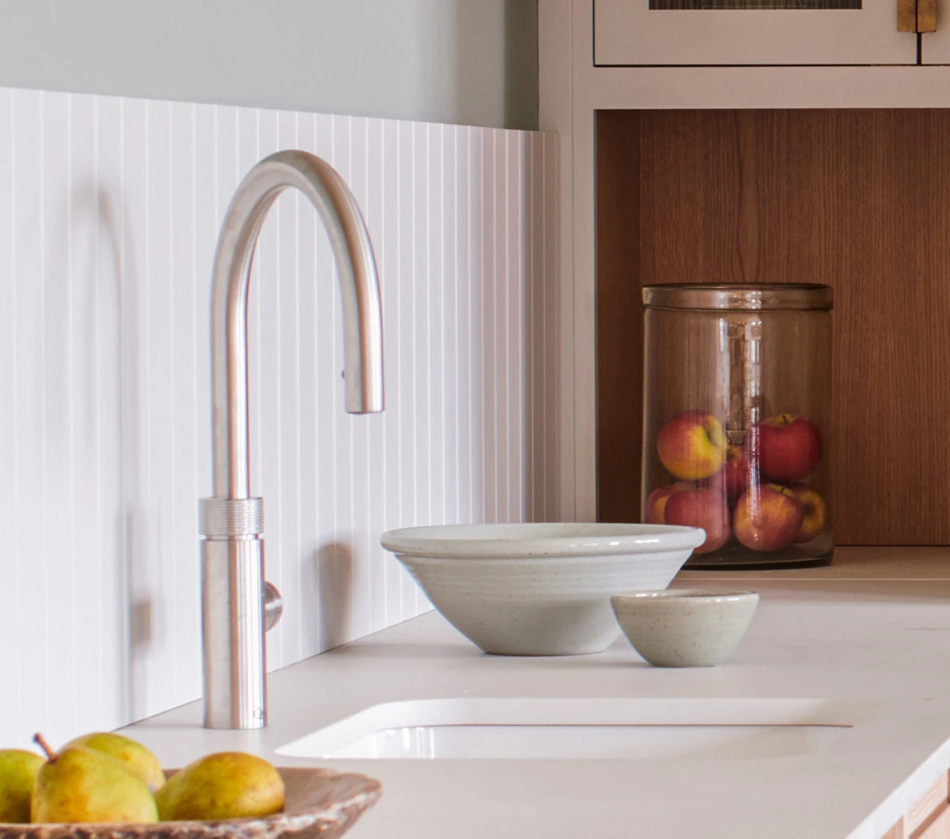 A modern kitchen countertop featuring a sleek silver faucet beside a white sink. Two ceramic bowls sit nearby, and a clear jar filled with apples is in the background. A small plate with pears is on the other side of the sink.