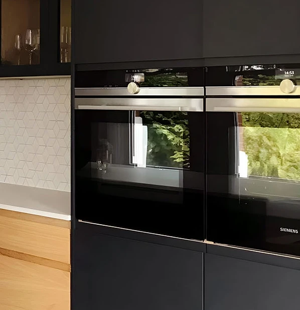 Modern kitchen with two black built-in ovens, sleek dark cabinetry, a light gray geometric-tiled backsplash, and light wood accents. Reflections of greenery visible in the oven doors.