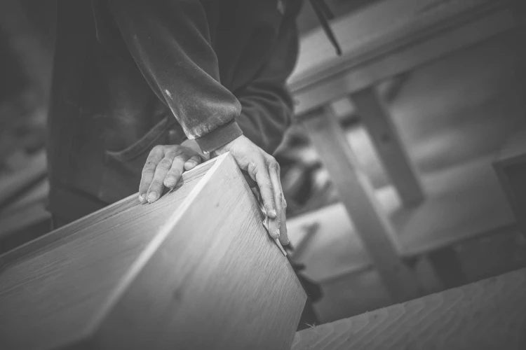 A person wearing a long-sleeve shirt uses both hands to work on a wooden piece in a workshop. The image is in black and white, emphasizing the focus on the craftsmanship and texture of the wood.