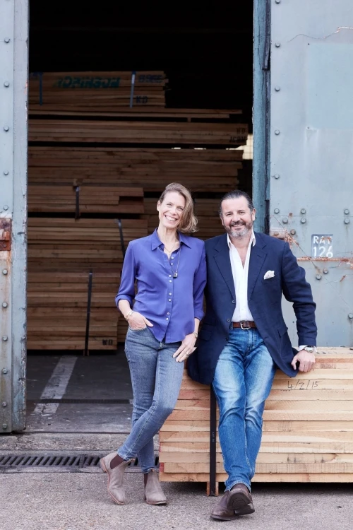 A woman and a man are leaning against a stack of wooden boards in front of a large open warehouse door. The woman is wearing a blue blouse and jeans, and the man is in a white shirt, blue blazer, and jeans. They are both smiling.