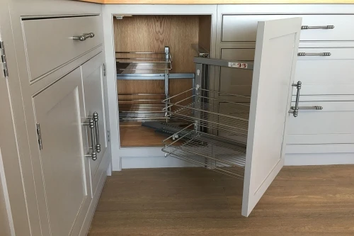 A white kitchen cabinet with a pull-out corner organizer is open, revealing metal wire shelving for storage. The cabinets have sleek metal handles, and the floor is light wood.