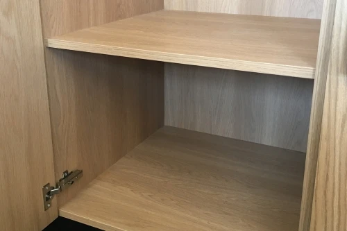 An open wooden cabinet with two empty shelves and a visible metal hinge on the door. The interior is made of light brown wood, and the cabinet appears new and clean