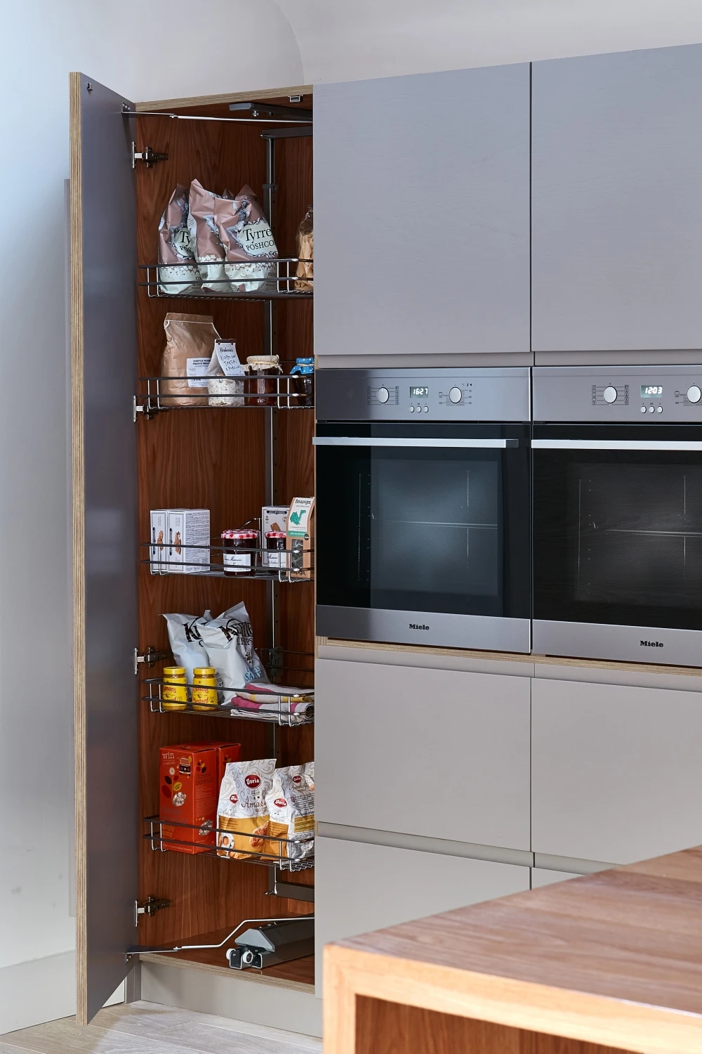 A modern kitchen with a tall, open pantry cupboard. The shelves are stocked with various food items and packages. Next to the pantry are two built-in stainless steel ovens. A wooden countertop is visible in the foreground.