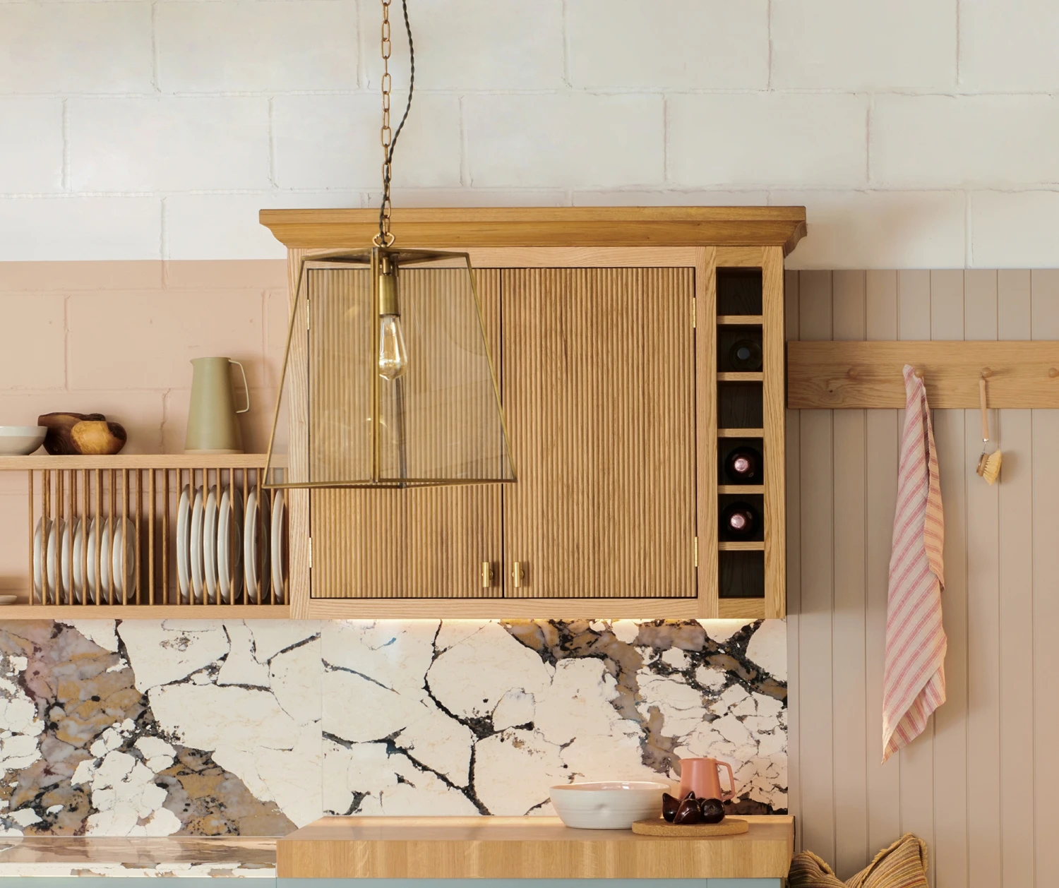 A cozy kitchen corner featuring light wooden cabinets, a marble backsplash, and a set of hanging cups. A modern pendant light hangs above a small countertop with kitchen items, and a striped towel is hung on the wall.