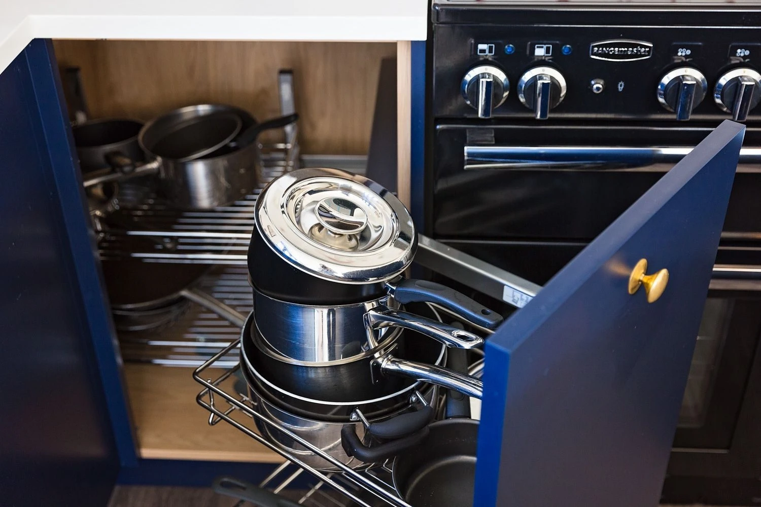 A corner kitchen cabinet with a pull-out shelf holds several stacked stainless steel pots and pans. The cabinet is partially open, displaying an organized storage setup next to a black oven with visible control knobs.