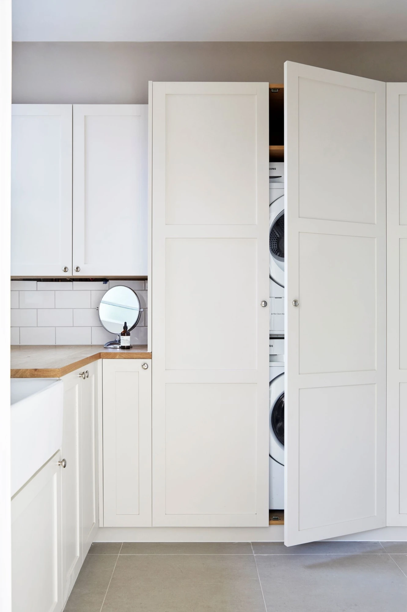 A modern laundry room with white cabinets. The double doors are open, revealing a stacked washer and dryer. The countertop has a small mirror and a bottle. The floor is tiled in a light gray color, and the backsplash features white subway tiles with grey grout.