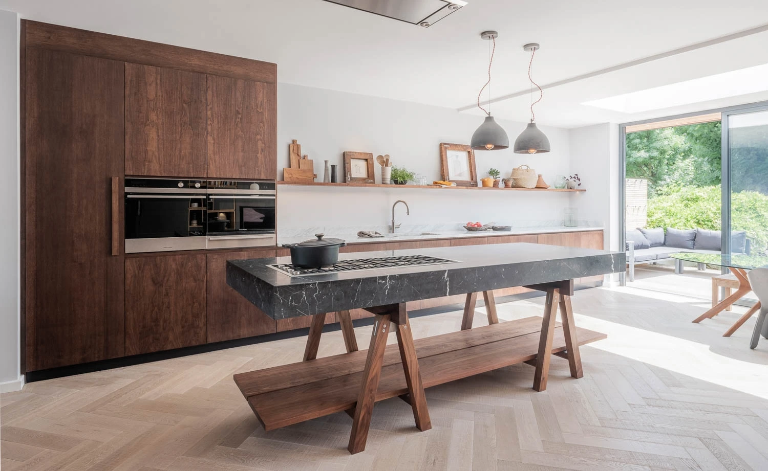 Modern kitchen with wooden cabinets, a marble island with a built-in stove and a pot, two pendant lights, and light wood herringbone flooring. A large window reveals a green outdoor view. Decorations and plants are on a long shelf above the counter.