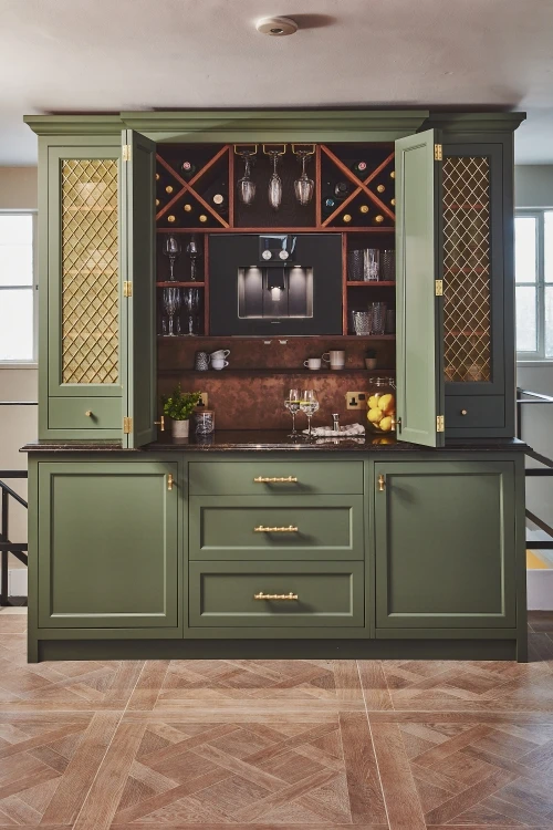 Elegant kitchen setup featuring green cabinets with mesh panel doors. Upper shelves hold wine glasses and bottles, while a small counter displays two filled wine glasses. Below, centered drawers and lower cabinets provide additional storage