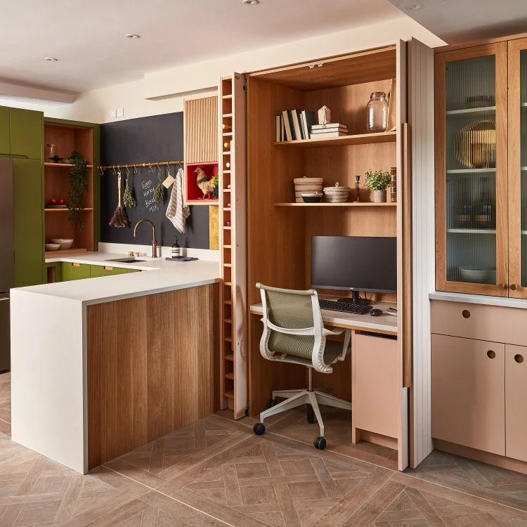 A modern kitchen with a white island features an integrated wooden workspace. The open cabinet reveals a desk with a computer and chair. Shelves hold decor items. The kitchen has green accents and a chalkboard wall with hanging utensils