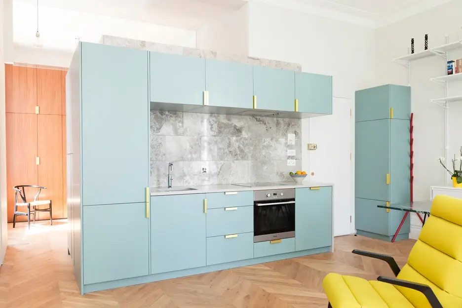 A modern kitchen with pastel blue cabinets and gold handles, featuring a built-in oven and marble backsplash. The room has herringbone wooden flooring and a yellow chair in the foreground.