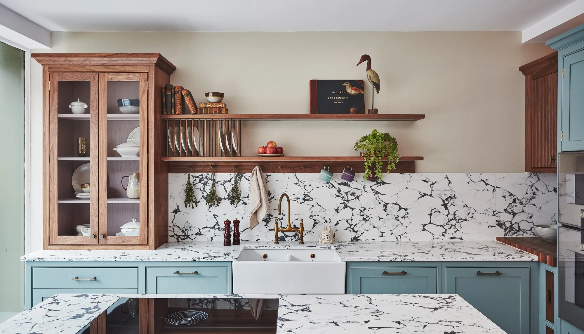 A cozy kitchen with a mix of styles. Light blue cabinets complement a marbled countertop and backsplash. A wooden shelf holds books and dishes, while a maroon island features a cutting board and red pot. Plants hang above the sink for decoration.