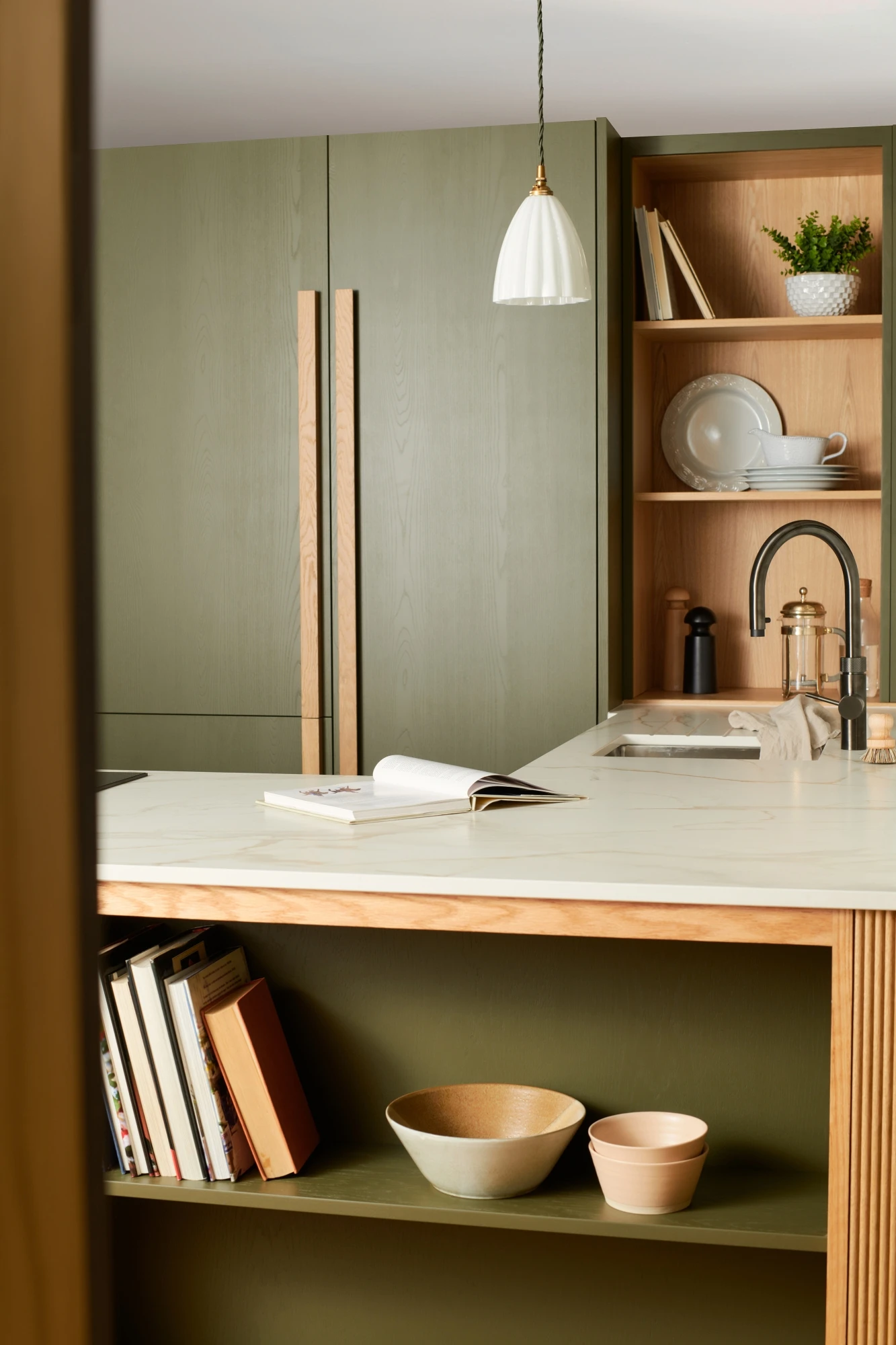 A modern kitchen with olive green cabinets, a marble countertop, and a built-in sink. Open shelves feature a plant, dishes, and bowls. A pendant light hangs above. The foreground includes a wooden bookshelf with various cookbooks and ceramic bowls