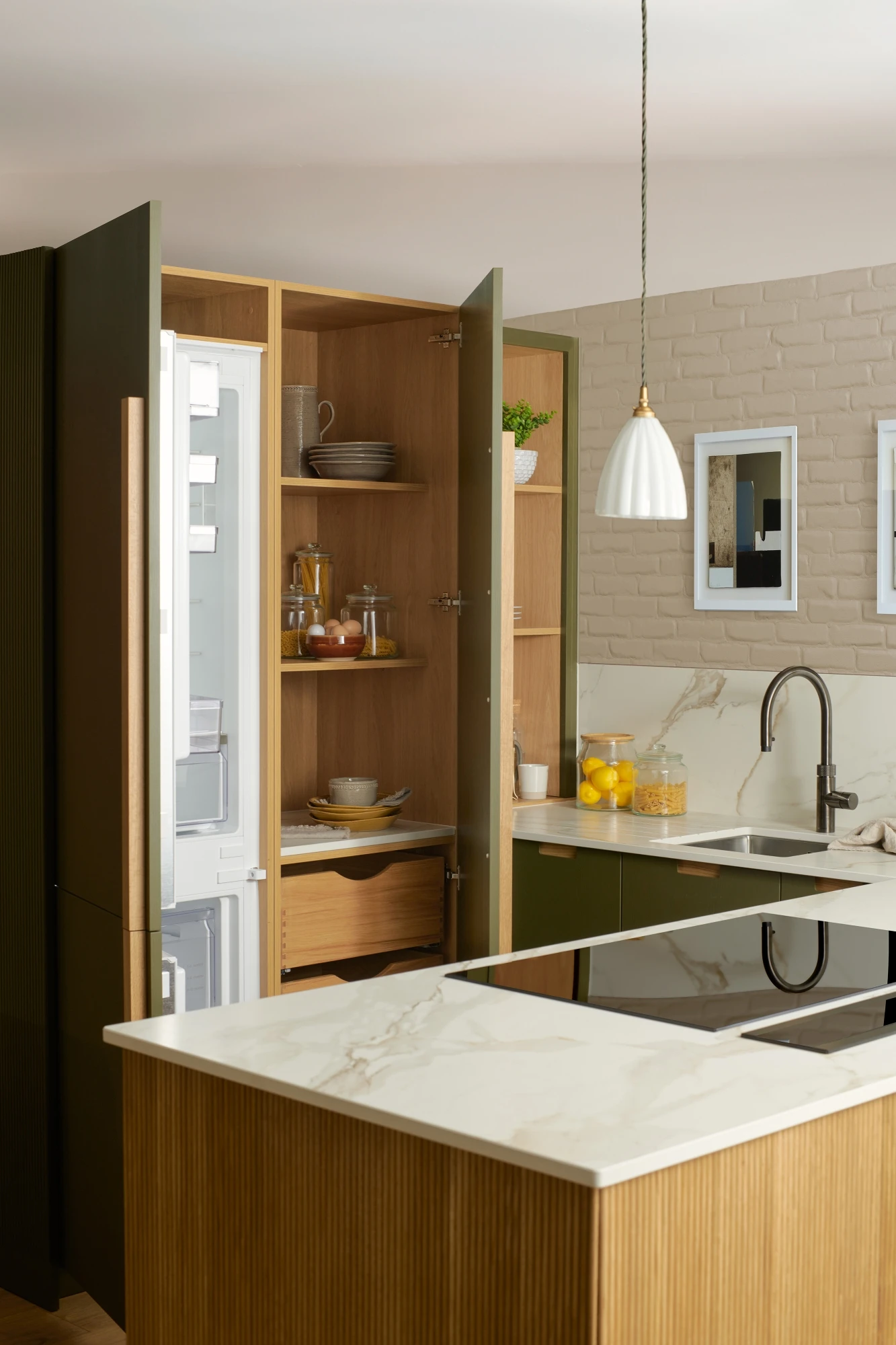 A modern kitchen with green cabinetry and a marble countertop. The kitchen island features an electric stovetop. There is a stainless steel sink with a gooseneck faucet, decorative items on shelves, and two framed pictures on a light brick wall.