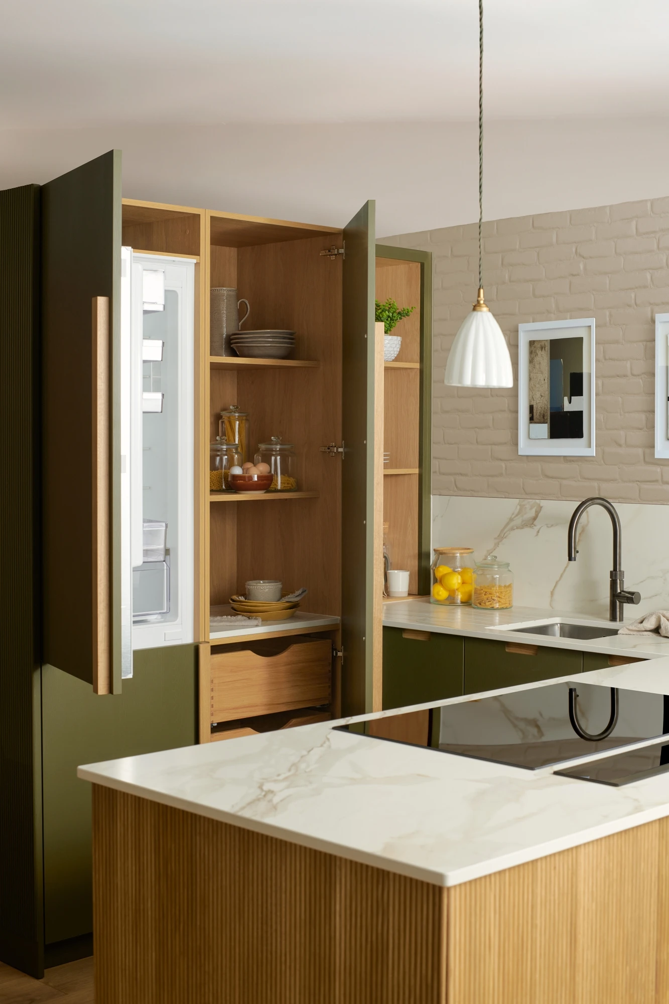 A modern kitchen with green cabinetry and a marble countertop. The kitchen island features an electric stovetop. There is a stainless steel sink with a gooseneck faucet, decorative items on shelves, and two framed pictures on a light brick wall.