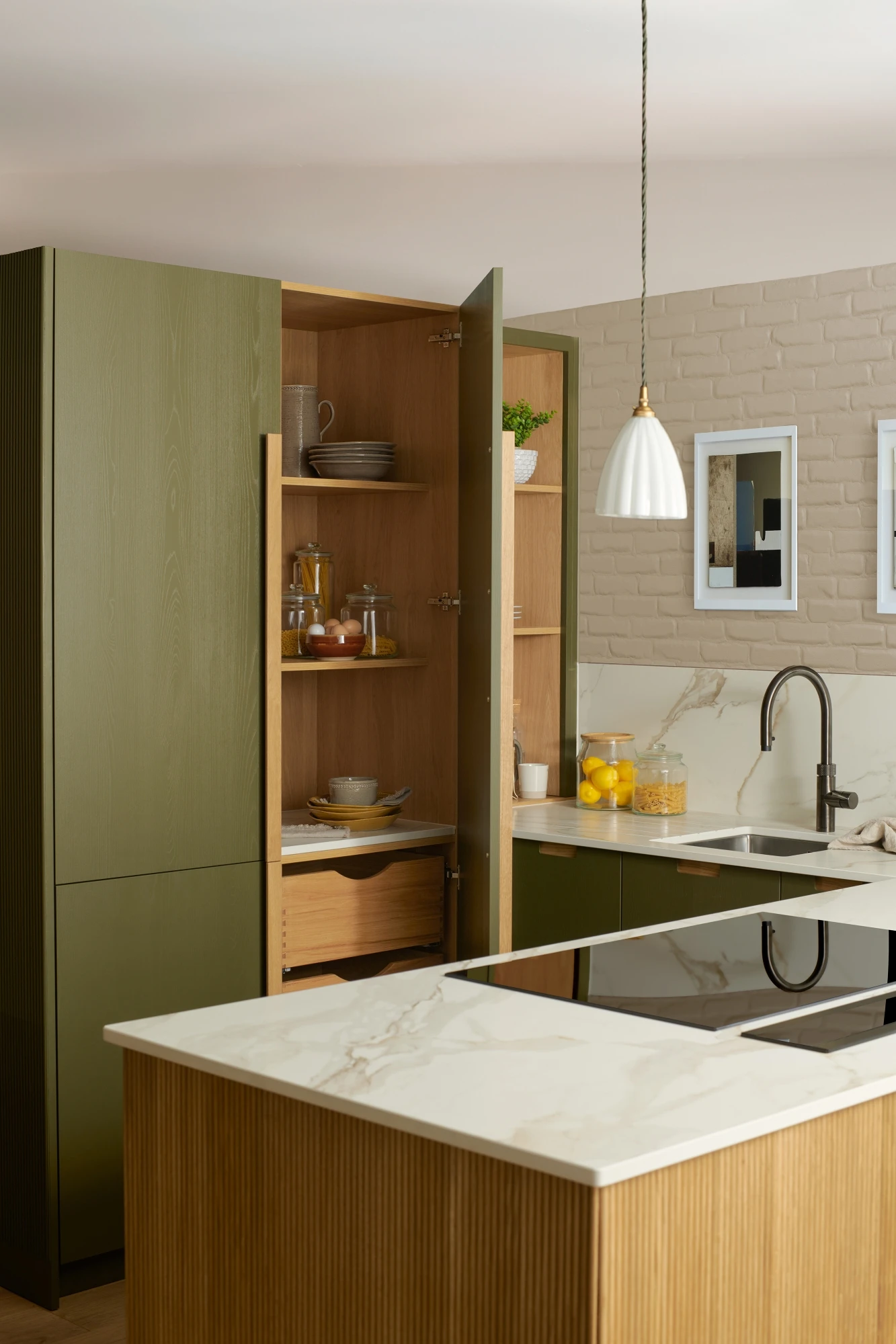 A modern kitchen with green cabinetry and a marble countertop. The kitchen island features an electric stovetop. There is a stainless steel sink with a gooseneck faucet, decorative items on shelves, and two framed pictures on a light brick wall.