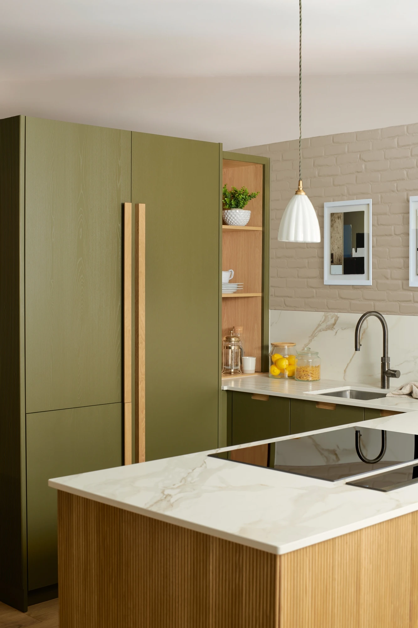 A modern kitchen with green cabinetry and a marble countertop. The kitchen island features an electric stovetop. There is a stainless steel sink with a gooseneck faucet, decorative items on shelves, and two framed pictures on a light brick wall.