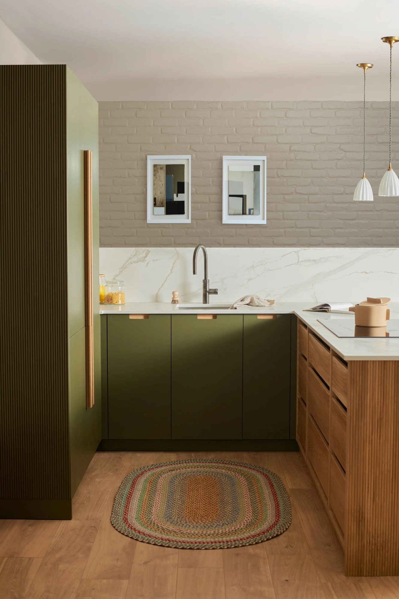 A modern kitchen with olive green and wood cabinetry, a white and gray marble countertop, and a light brown textured rug. Two pendant lights hang over an island. Beige brick and marble backsplash add contrast. Two framed pictures are on the wall.