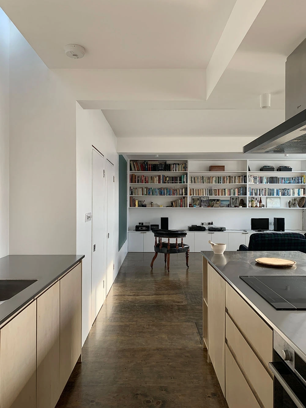 Minimalist open-plan kitchen and living area with wooden floors. In the foreground, a kitchen island with countertops and cabinetry. In the background, a wall filled with bookshelves, a round table, and a cozy seating area. Natural light fills the space.