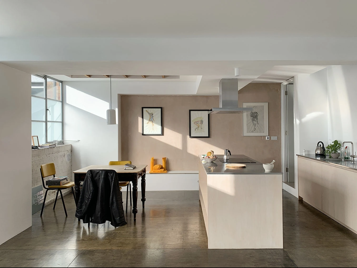 A modern kitchen with natural light streaming through large windows. It features a central island with an overhead hood, a dining table with chairs, wall art, and sleek cabinets. There's a jacket draped over one chair. The floor is polished concrete.