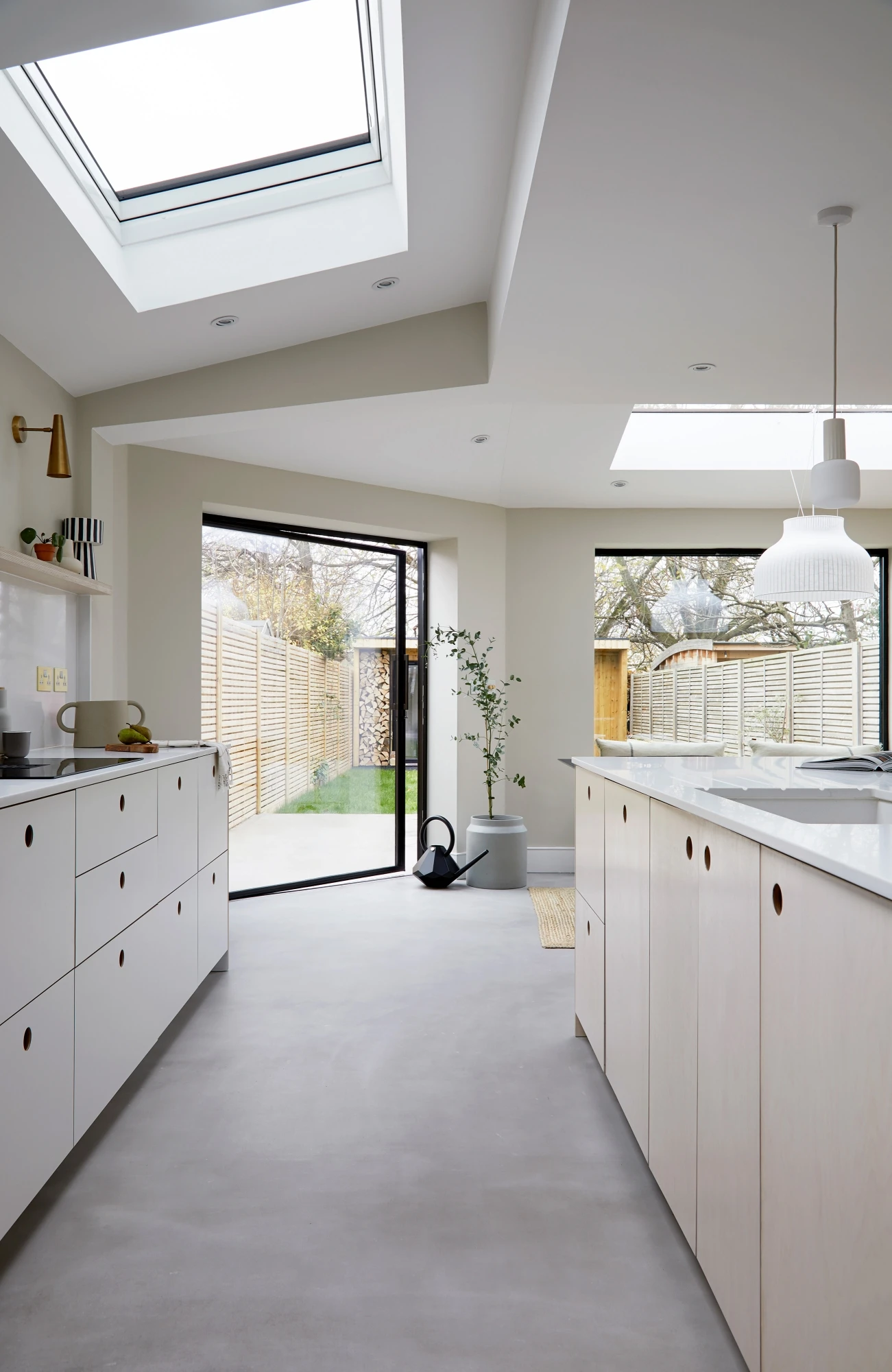 Modern kitchen with white cabinets, minimalist design, and large skylights. The room features an island, pendant lighting, and a potted plant. A sliding glass door leads to a fenced backyard. The space is filled with natural light.