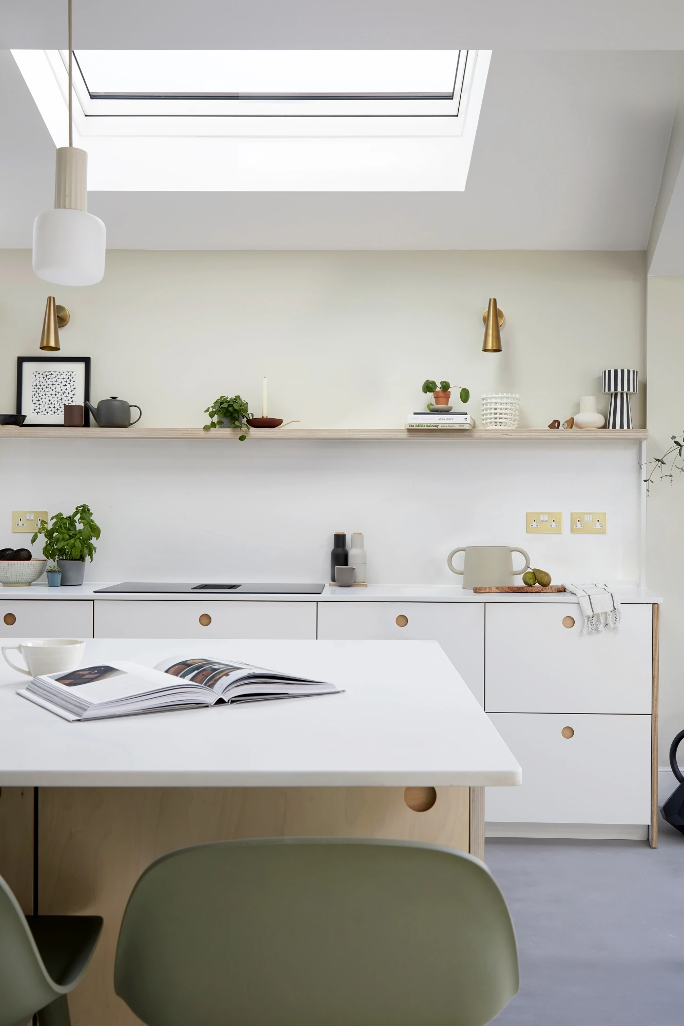 Modern kitchen interior with white cabinetry and a minimalist aesthetic. An open book is placed on a central island countertop. Shelves display small plants, kitchenware, and decor items. A skylight above illuminates the room with natural light.