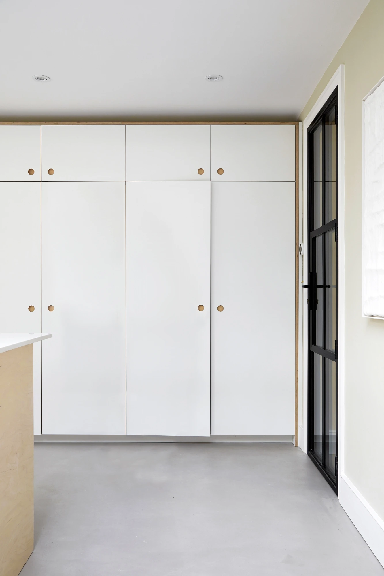 A minimalist kitchen space with clean white cabinets and a light-colored countertop. The cabinets feature wooden knobs, and there is a black-framed glass door on the right. The walls are painted in a light beige tone, and natural light brightens the room.