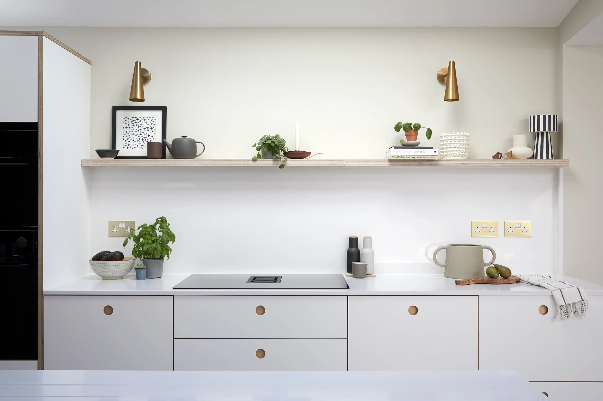 Modern kitchen featuring white cabinetry with wooden knobs, an induction stove, and a smooth white countertop. In the background, a wooden floating shelf displays various decorative items, plants, and books. Vertical brass lights are mounted on the wall.