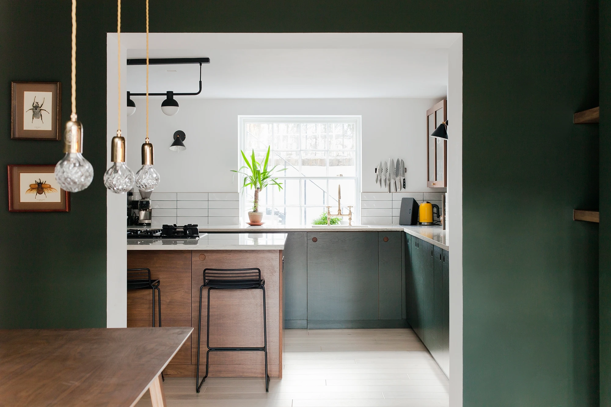 A modern kitchen with dark green walls and white ceiling. It features wooden cabinetry, a marble countertop, bar stools, and a gas stove. A large window provides natural light. Three pendant lights with clear glass shades hang over the island, and insect art decorates the wall.