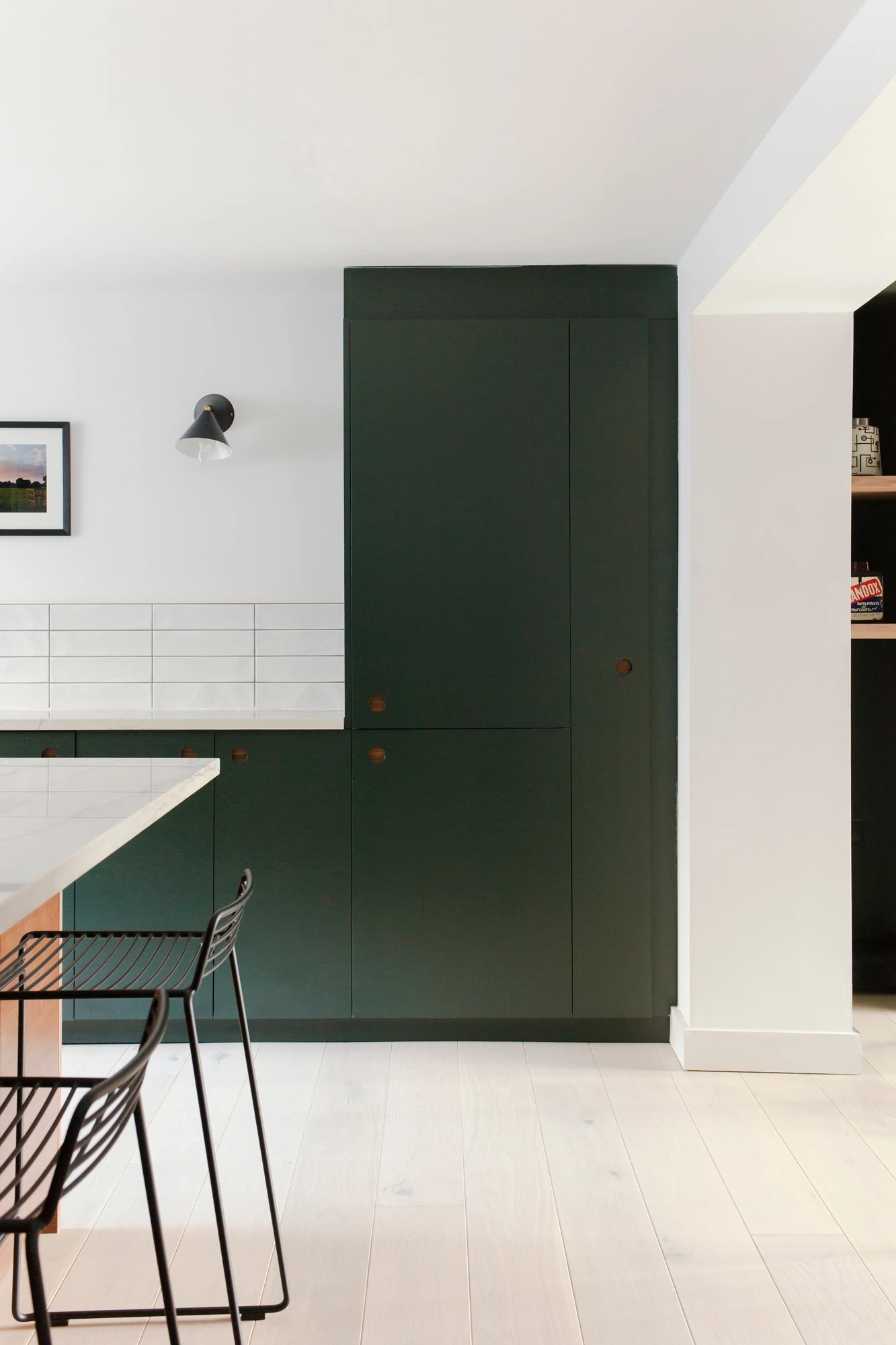 Modern kitchen with dark green cabinets and white tile backsplash. A black sconce light is mounted above the backsplash, and a part of the kitchen island with a light-colored countertop and black wire bar stools is visible. Walls and floors are light-colored.