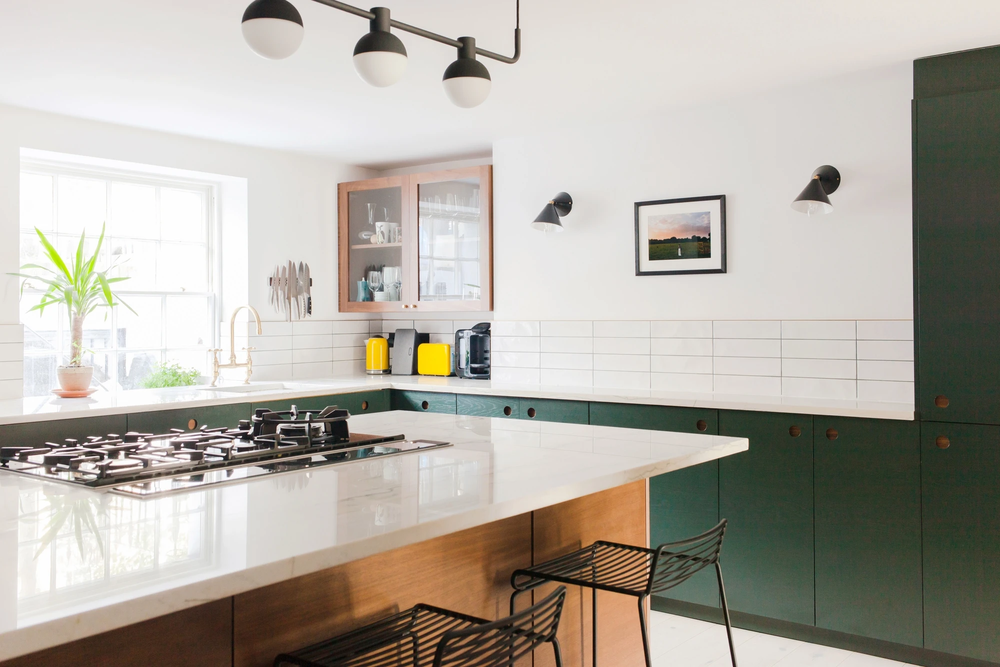 A modern kitchen with a large central island featuring a gas stove. Surrounding white cabinets are topped with green cabinets and a white tiled backsplash. Black barstools sit by the island. A large window and wall-mounted lights provide ample lighting.