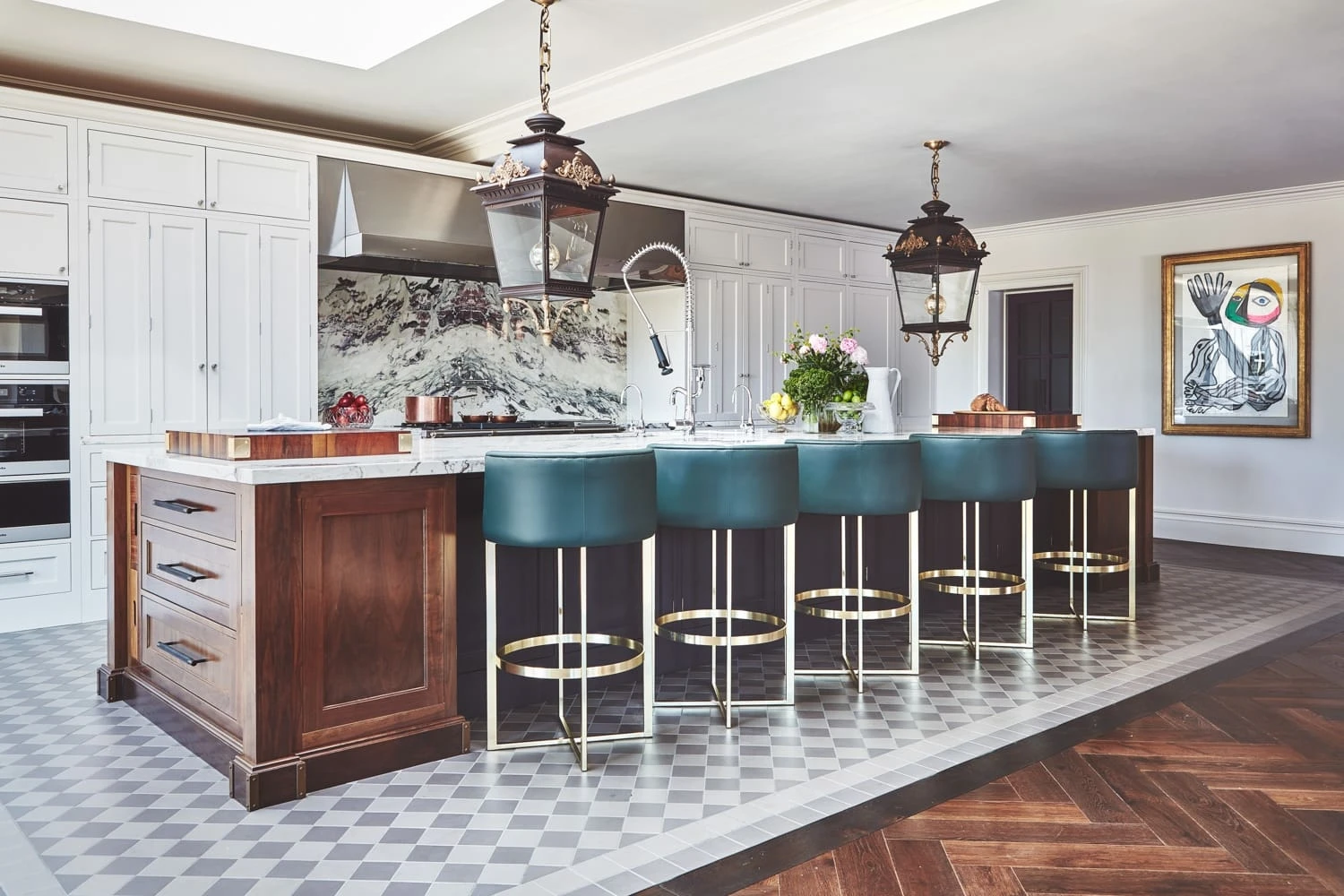 A modern kitchen with a large island featuring a wooden base, a marble countertop, and five green bar stools with gold accents. White cabinetry and a colorful backsplash dominate the background. Hanging pendant lights illuminate the space, with a piece of artwork on the wall.