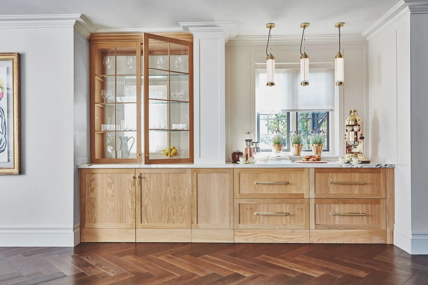 A modern kitchen area with wooden cabinetry and glass-front cabinets displaying glassware. A countertop features potted plants, glass containers, and a large coffee machine. Three pendant lights hang above. Dark hardwood flooring contrasts with the light décor.