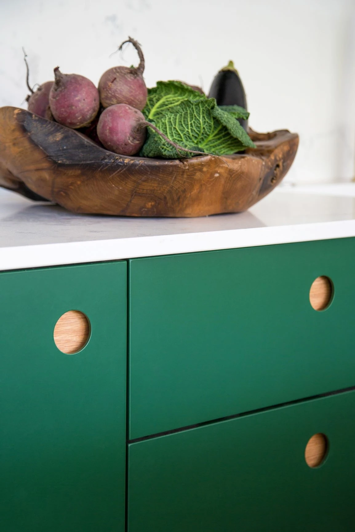 A wooden bowl with beets, cabbage, and an eggplant sits on a white countertop above green cabinets with circular wooden handles.