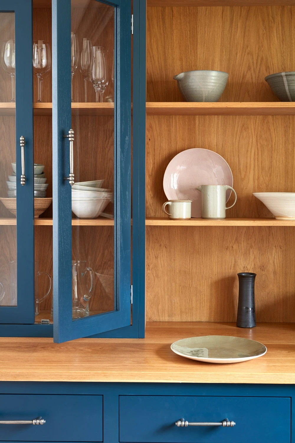 A wooden kitchen cabinet with open shelves displays various ceramic dishes and glassware. The lower cabinet is painted blue, featuring polished silver handles. A black vase and a large plate sit on the wooden countertop.