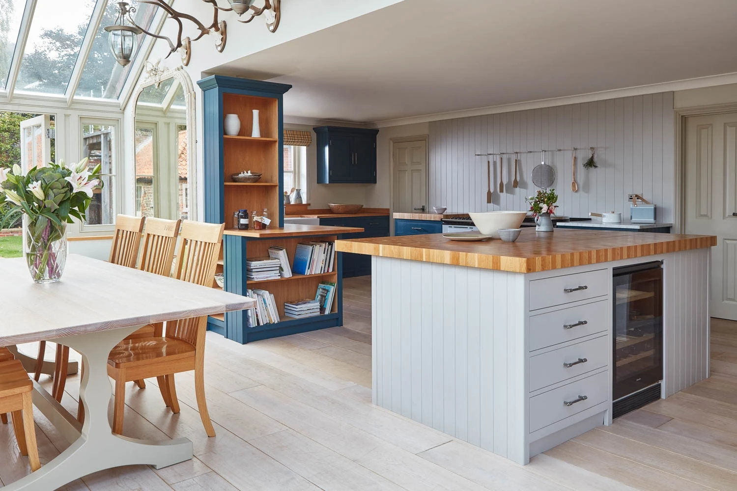 A spacious kitchen with light wood flooring and a dining area featuring wooden chairs and a white table. The kitchen has two-tone cabinets, one island with shelves and a wine cooler, and another with open shelving. Large windows brighten the space.