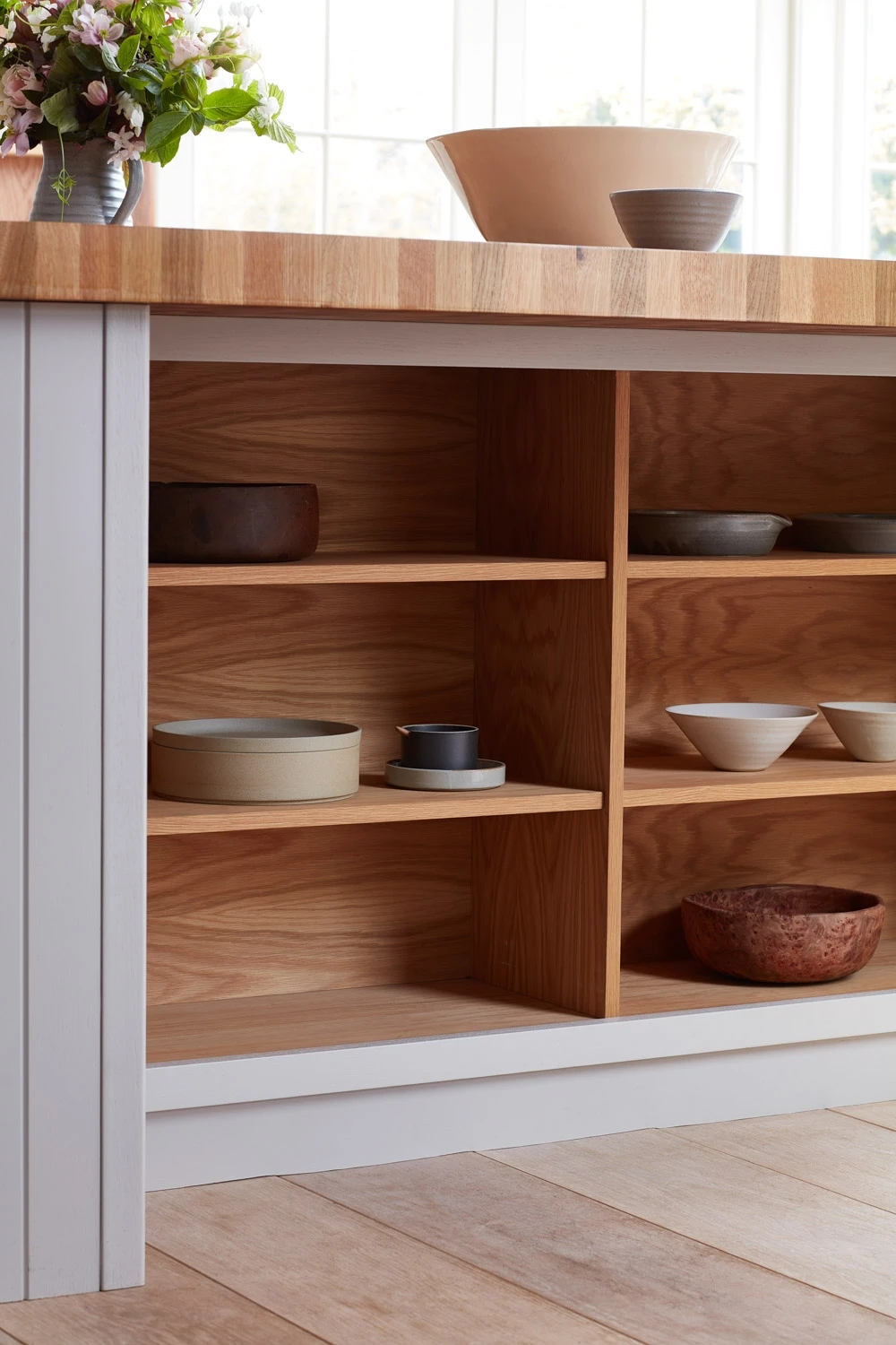 A wooden kitchen island with open shelves displaying various ceramic bowls and dishes, under a butcher block countertop. A vase with pink flowers sits on the countertop by a sunlit window