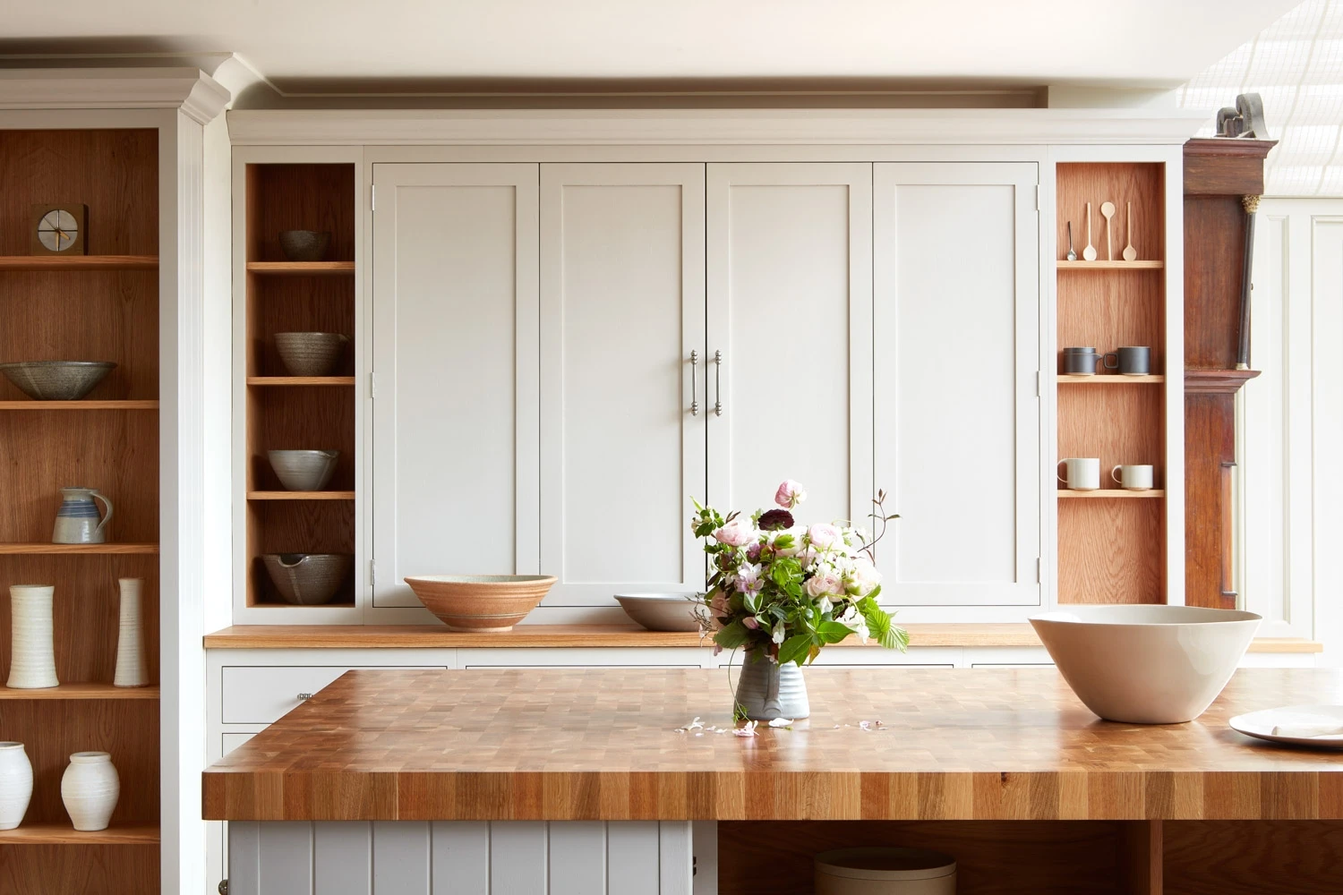 A modern kitchen with white cabinets and wooden open shelves displaying various bowls and vases. A wooden island stands in the center with a vase of fresh flowers and large white bowls on top. The room is bright and well-organized