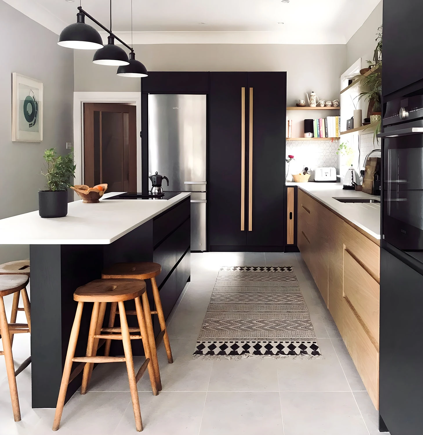 A modern kitchen with black cabinets, light wooden drawers, and stainless steel appliances. It features an island with bar stools, pendant lights overhead, and a patterned rug on the light tile floor. Shelves with various items and plants are on the walls.