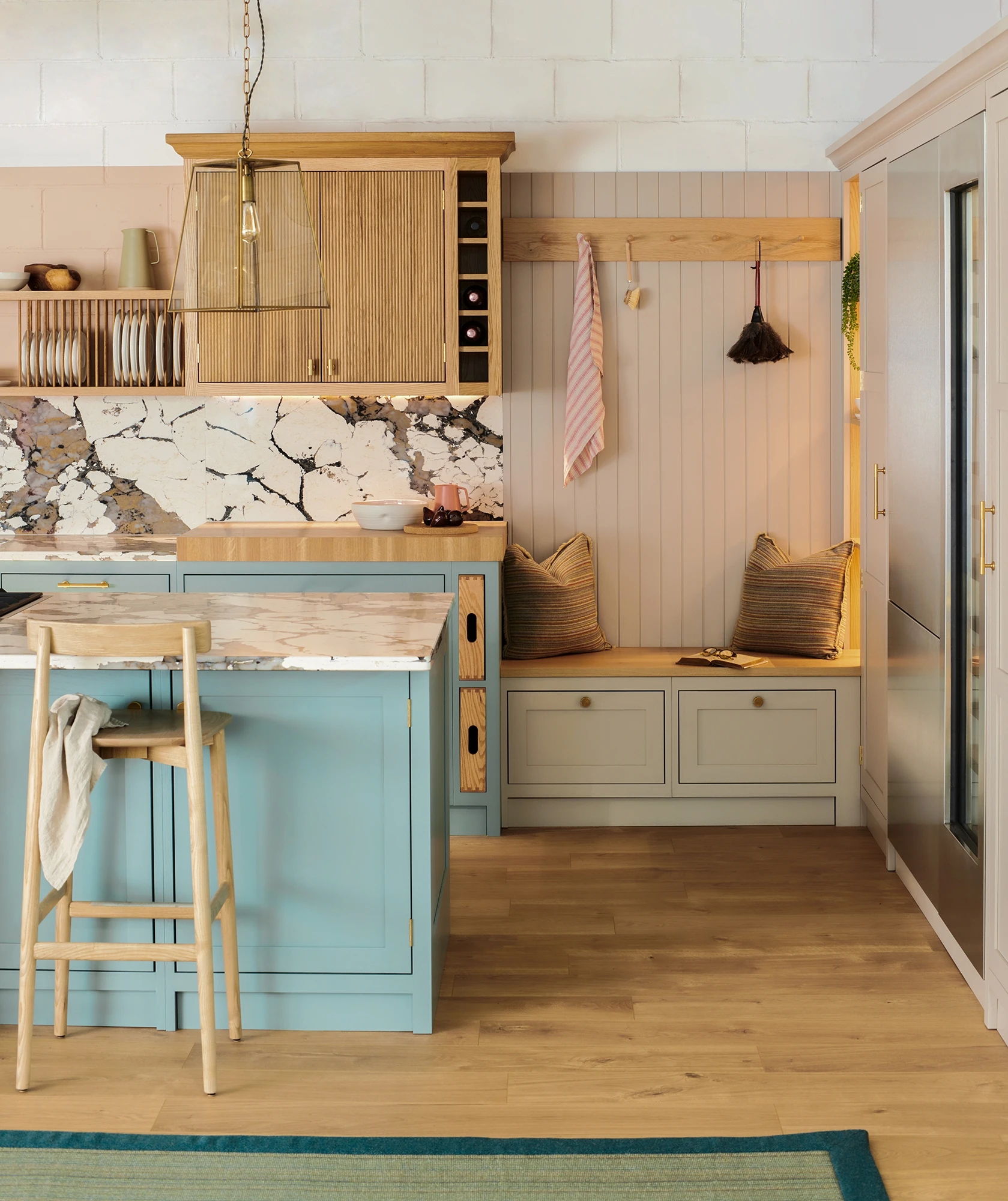 A cozy kitchen features light wooden cabinetry, a marble-topped island painted in pastel blue with a wooden stool, and a built-in seating nook with pillows and hanging hooks. The floor is hardwood, and there are decorative accents like a stylish pendant light.