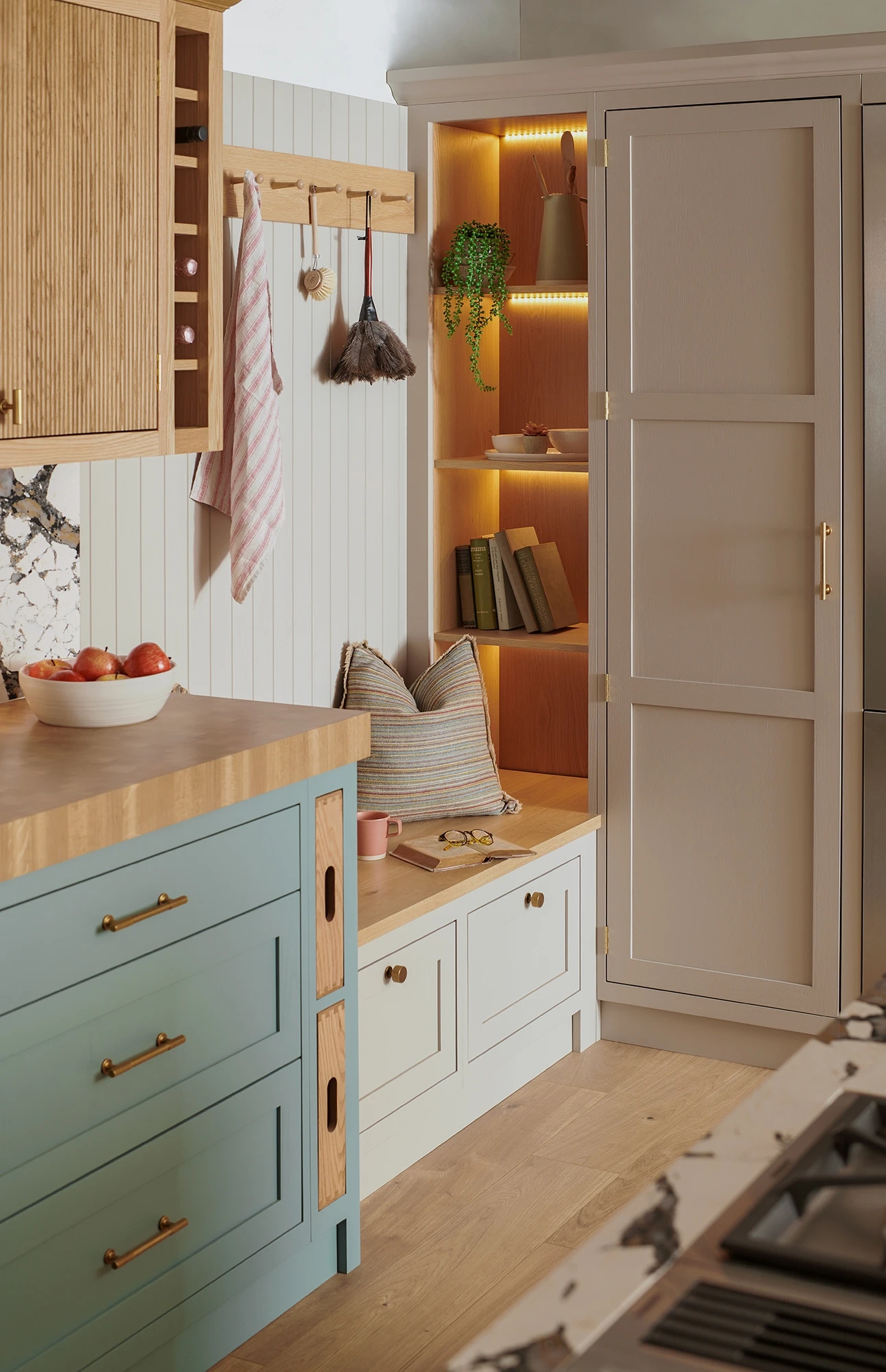 A cozy kitchen corner featuring a built-in reading nook with shelves holding books and a plant. The nook is illuminated by warm LED lights and has a cushioned bench. The kitchen has light blue and wooden cabinets, a bowl of tomatoes, and stylish decor.