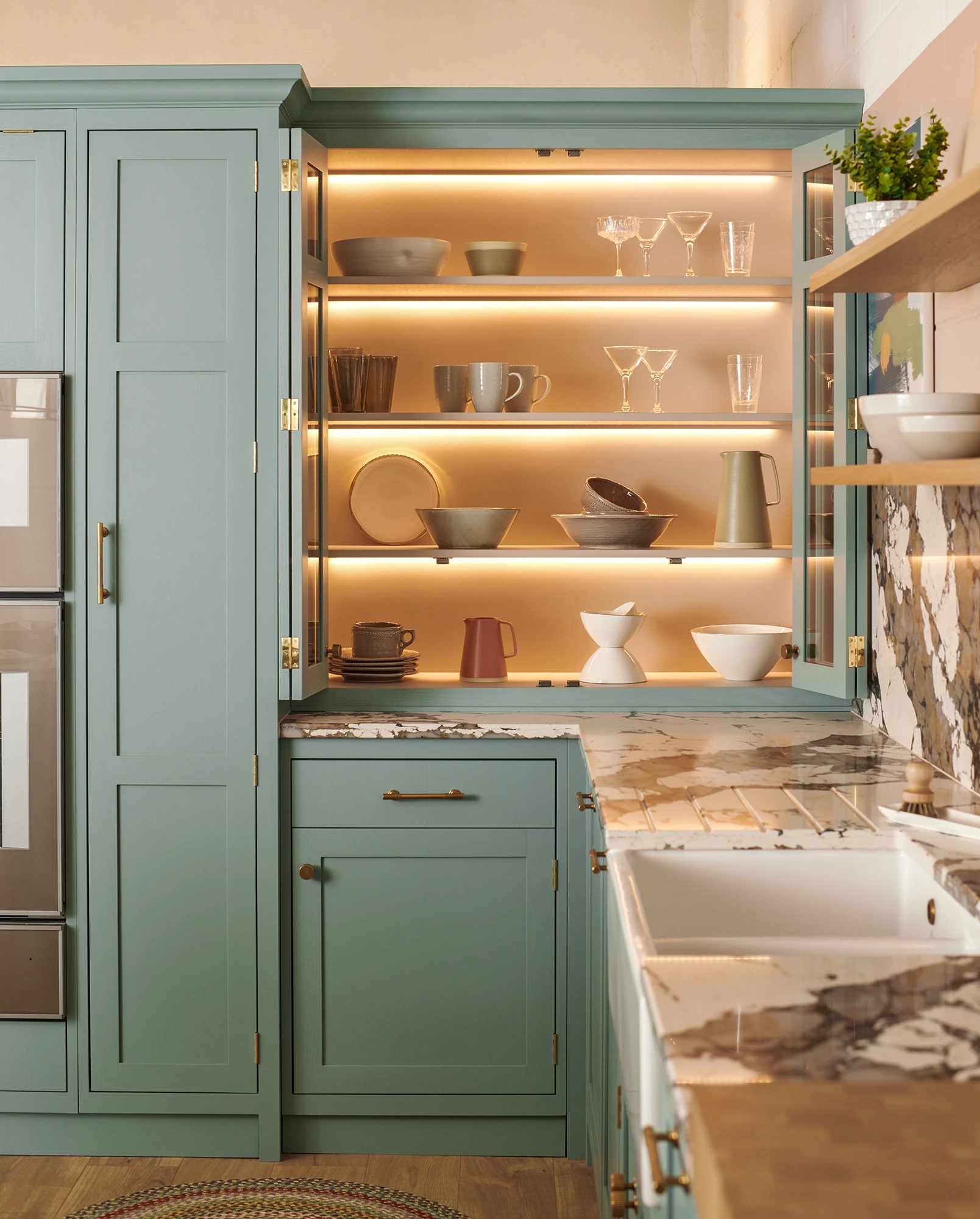 A modern kitchen with sage green cabinets, open shelving displaying neatly arranged dishware and glassware with soft under-cabinet lighting. The countertops are marble, and there's a farmhouse sink with a brass faucet. Shelves hold bowls, mugs, and glasses