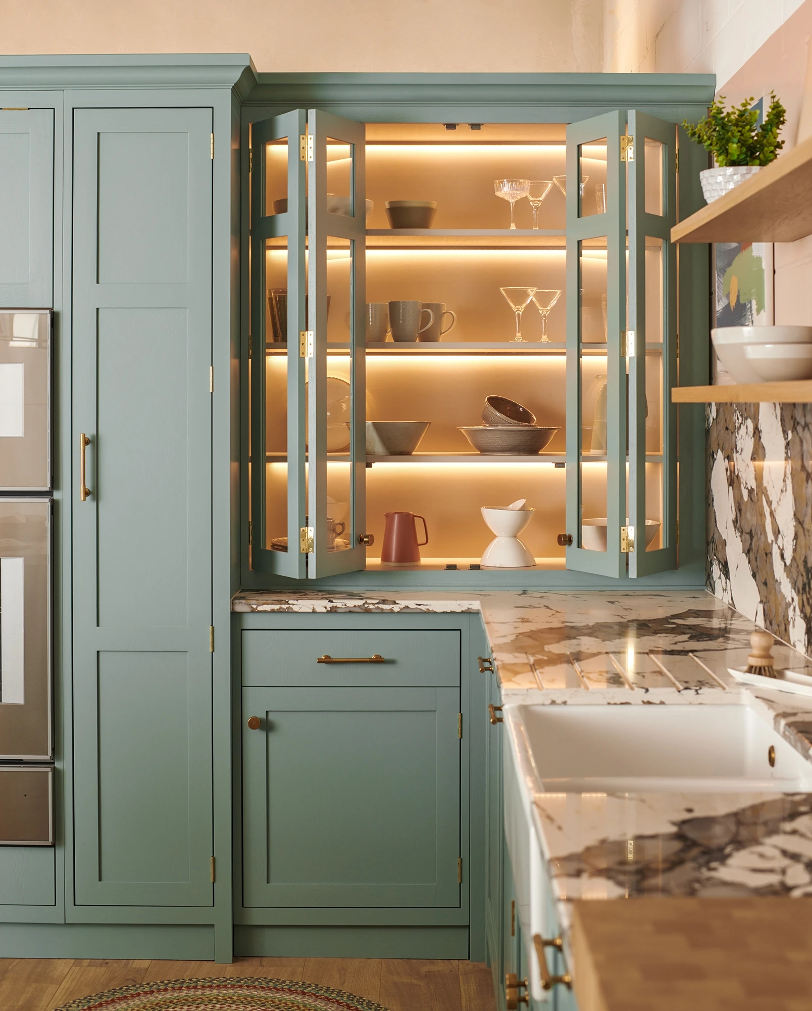 A modern kitchen with sage green cabinets, open shelving displaying neatly arranged dishware and glassware with soft under-cabinet lighting. The countertops are marble, and there's a farmhouse sink with a brass faucet. Shelves hold bowls, mugs, and glasses