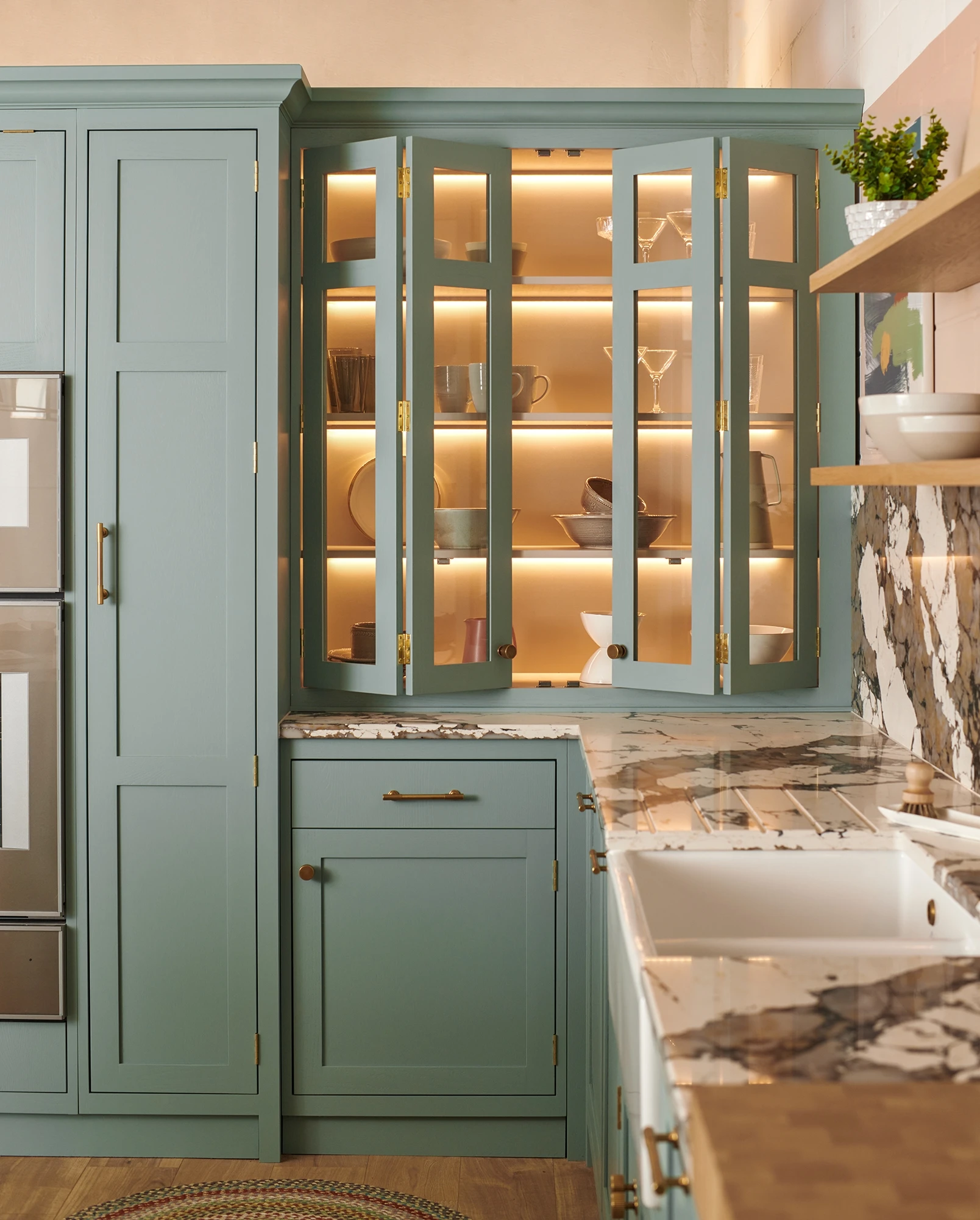 A modern kitchen with sage green cabinets, open shelving displaying neatly arranged dishware and glassware with soft under-cabinet lighting. The countertops are marble, and there's a farmhouse sink with a brass faucet. Shelves hold bowls, mugs, and glasses