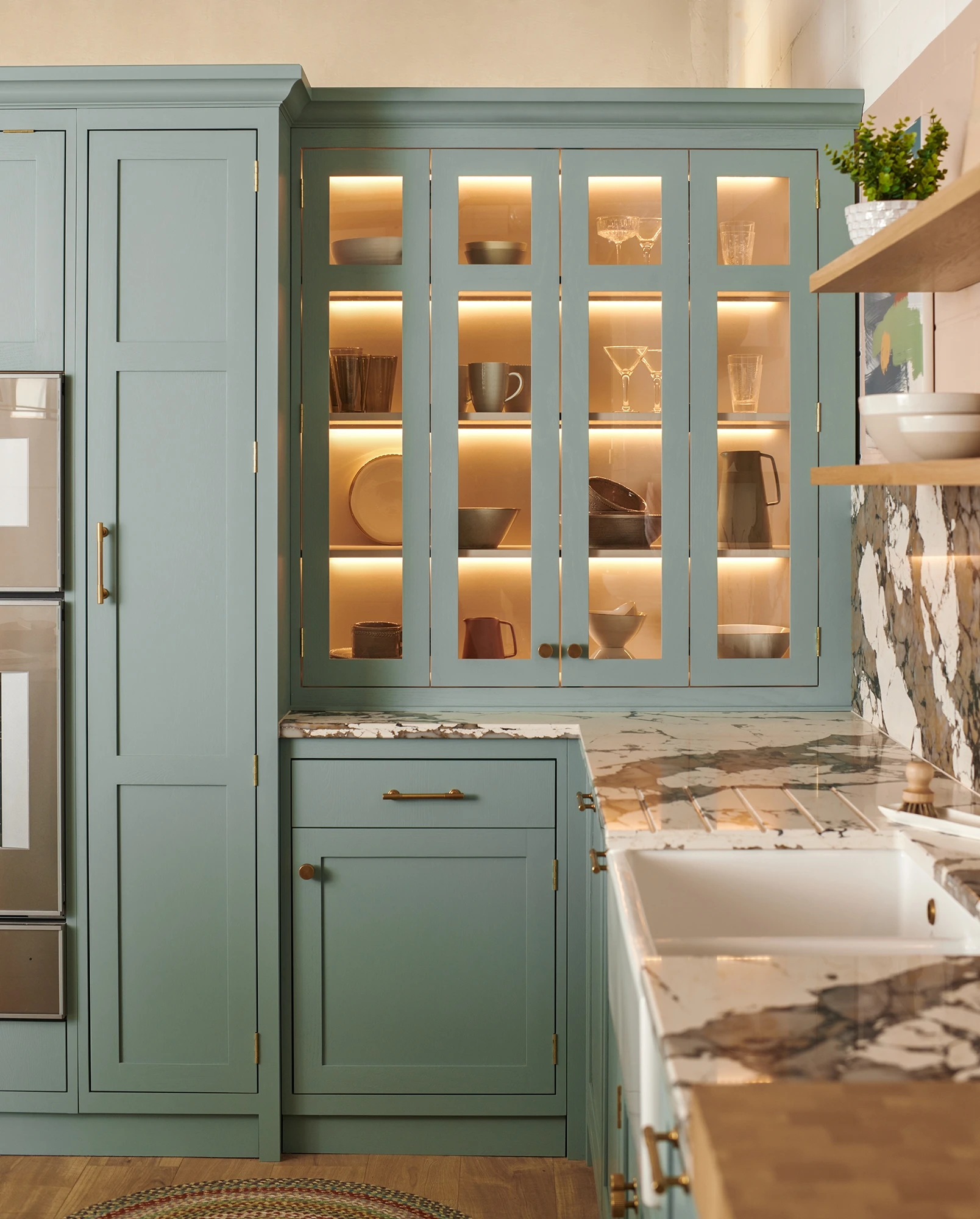 A modern kitchen with sage green cabinets, open shelving displaying neatly arranged dishware and glassware with soft under-cabinet lighting. The countertops are marble, and there's a farmhouse sink with a brass faucet. Shelves hold bowls, mugs, and glasses